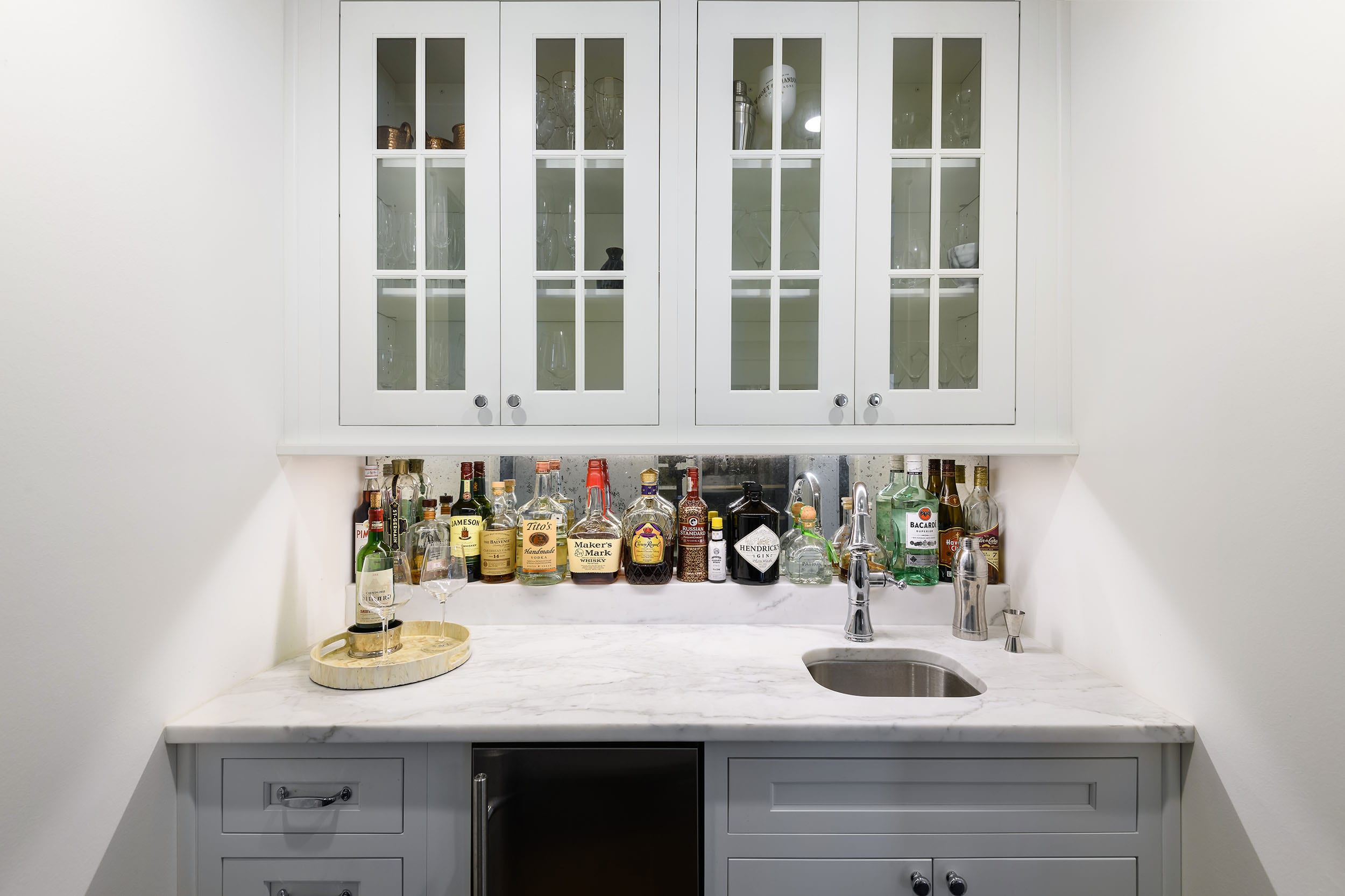 A stylish home bar with various liquors on a marble countertop under white cabinets.