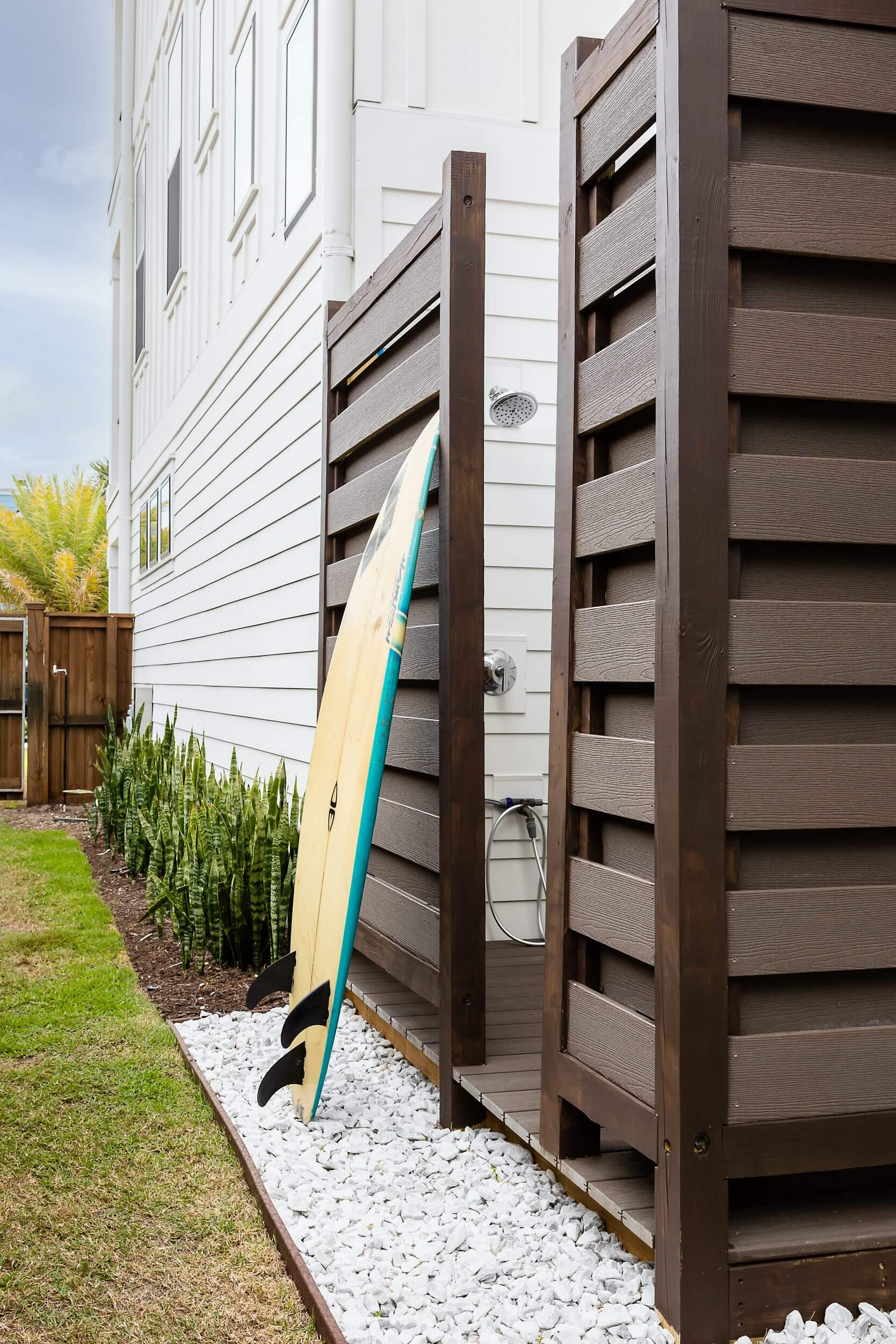 Surfboard leaning against an outdoor shower beside a white house.